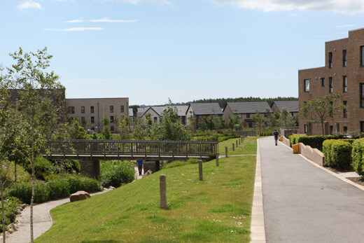 A paved walkway runs through a residential area with modern brick apartment buildings on the right and a grassy area with a wooden bridge.